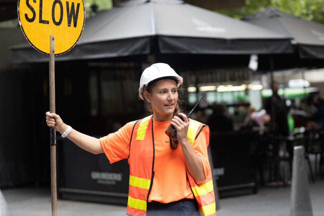 A construction worker holds a "SLOW" sign and uses a walkie-talkie